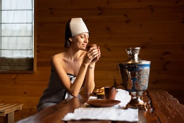 Young woman in a sauna with a cap on her head sits at a table and drinks herbal tea, enjoying a wellness day